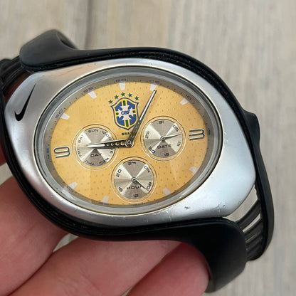 Nike Triax “Brasil” Swift 3i WR0091 analog watch with black rubber straps and a silver-tone case featuring a black Nike Swoosh. The yellow metallic dial has a recessed dotted texture, a Brazil crest at the top, three silver subdials for day, date, and hour, silver hands, and a green seconds hand. Seconds markings are printed on the inner edge of the case and are visible only at an angle. Sport-focused Triax case design with water-resistant construction.