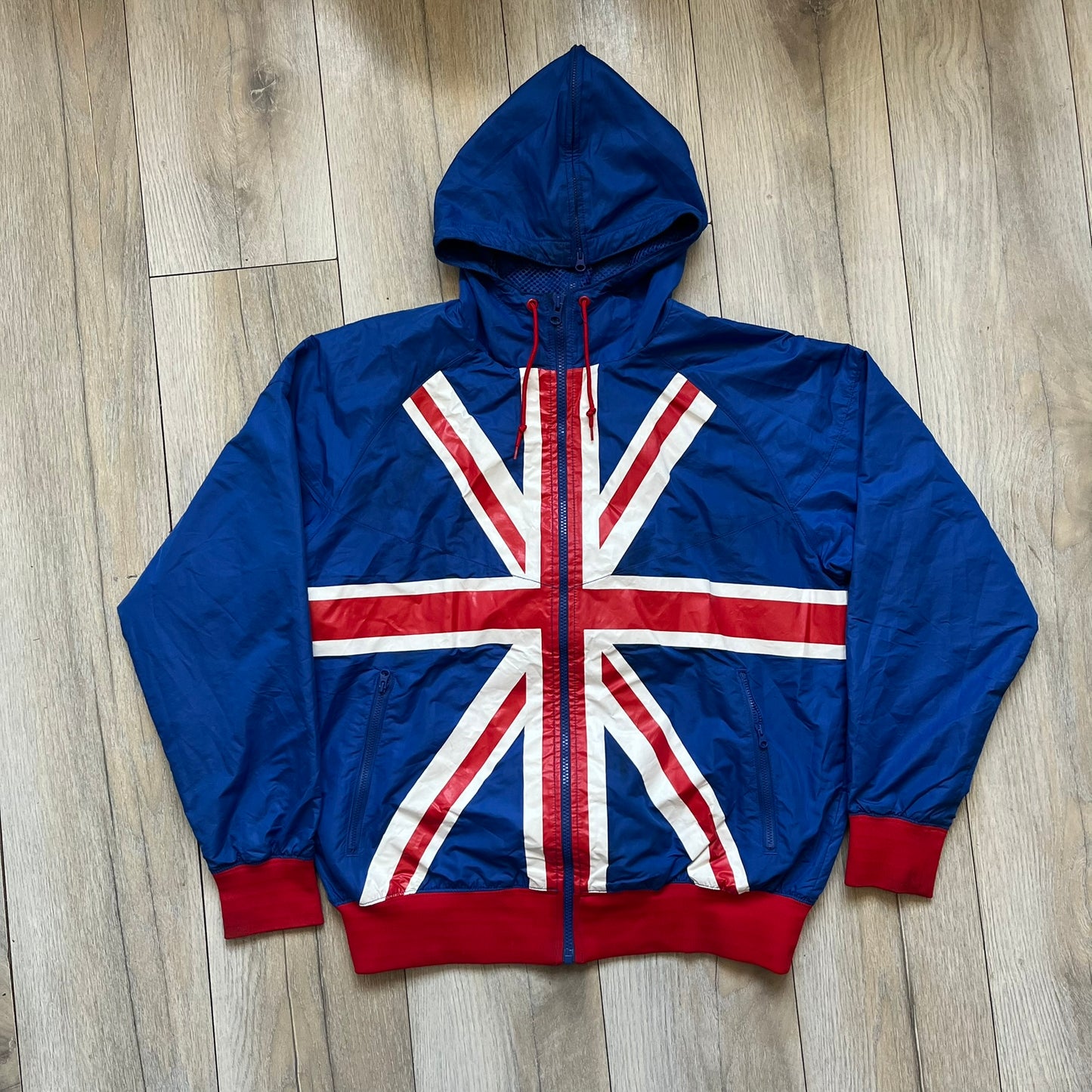 Nike UK Olympics 2008 Split Windbreaker designed by Jarrett Reynolds. Features a blue base with Union Jack-inspired red and white stripes on the front, representing Great Britain. Dual side zippers allow the jacket to be split in half and combined with other pieces from the collection, with a subtle United Kingdom flags inside the pocket.