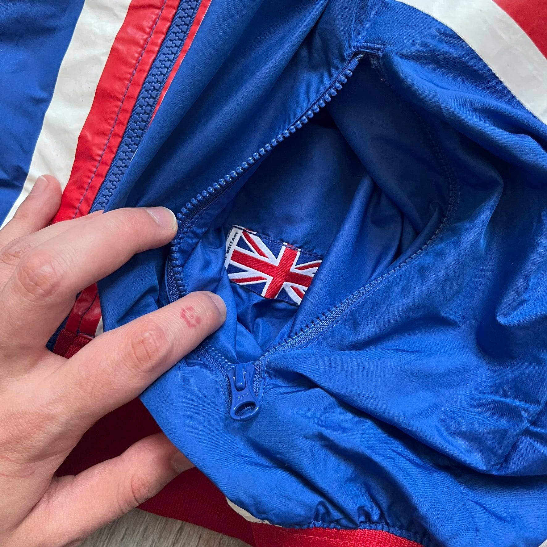 Nike UK Olympics 2008 Split Windbreaker designed by Jarrett Reynolds. Features a blue base with Union Jack-inspired red and white stripes on the front, representing Great Britain. Dual side zippers allow the jacket to be split in half and combined with other pieces from the collection, with a subtle United Kingdom flags inside the pocket.