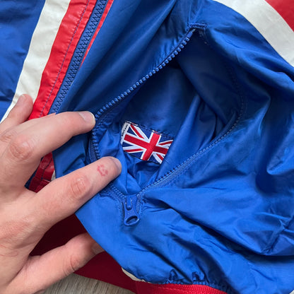 Nike UK Olympics 2008 Split Windbreaker designed by Jarrett Reynolds. Features a blue base with Union Jack-inspired red and white stripes on the front, representing Great Britain. Dual side zippers allow the jacket to be split in half and combined with other pieces from the collection, with a subtle United Kingdom flags inside the pocket.