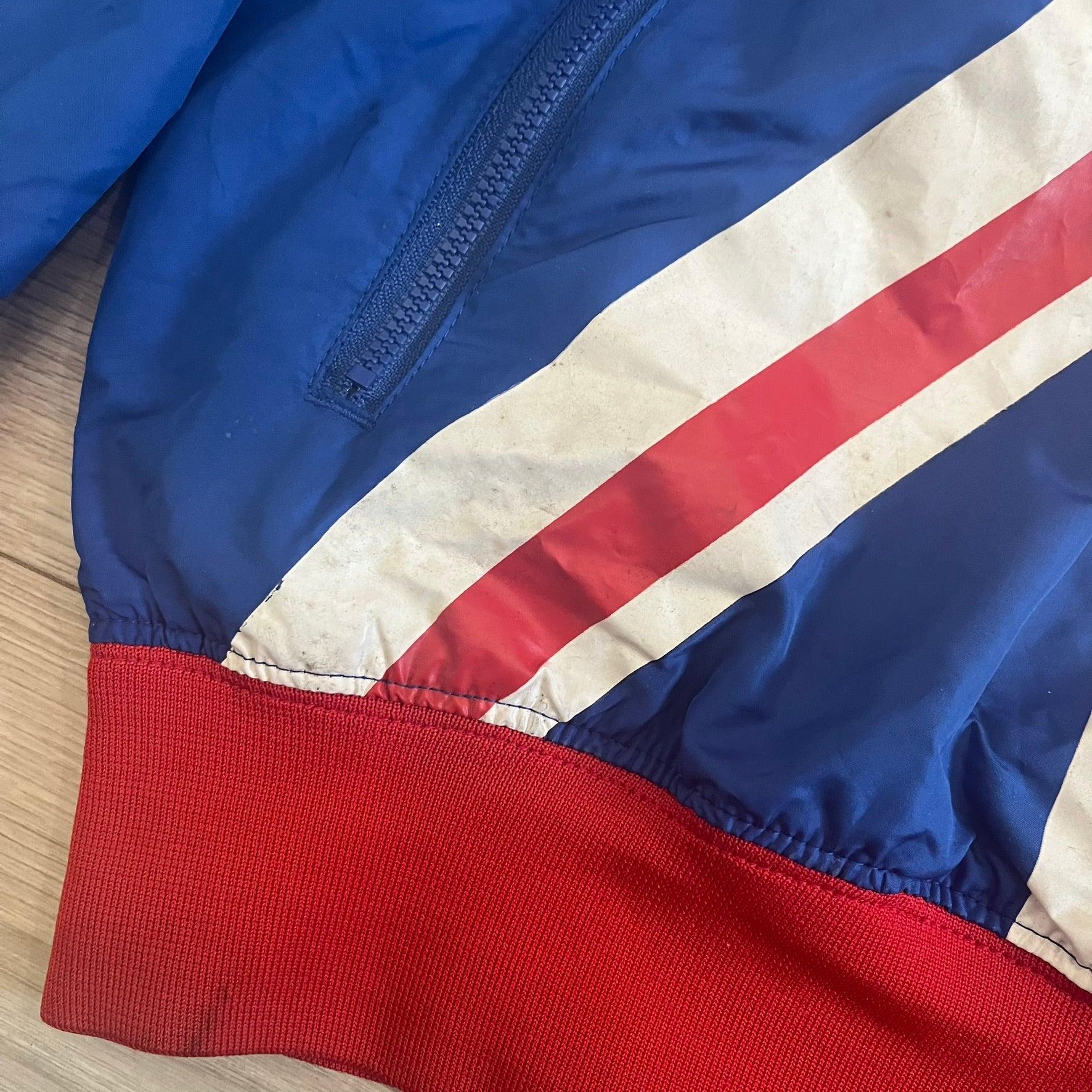 Nike UK Olympics 2008 Split Windbreaker designed by Jarrett Reynolds. Features a blue base with Union Jack-inspired red and white stripes on the front, representing Great Britain. Dual side zippers allow the jacket to be split in half and combined with other pieces from the collection, with a subtle United Kingdom flags inside the pocket.