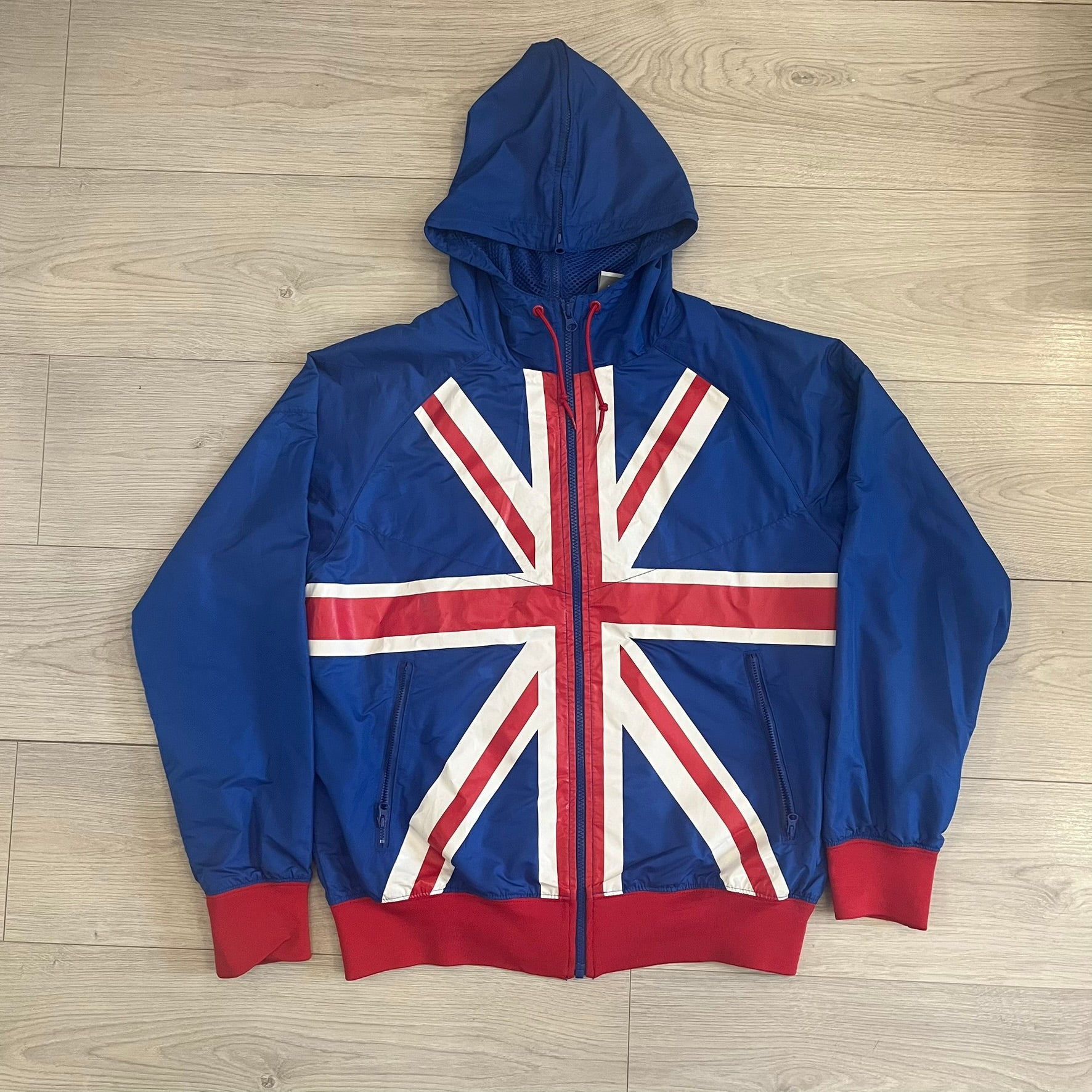 Nike UK Olympics 2008 Split Windbreaker designed by Jarrett Reynolds. Features a blue base with Union Jack-inspired red and white stripes on the front, representing Great Britain. Dual side zippers allow the jacket to be split in half and combined with other pieces from the collection, with a subtle United Kingdom flags inside the pocket.