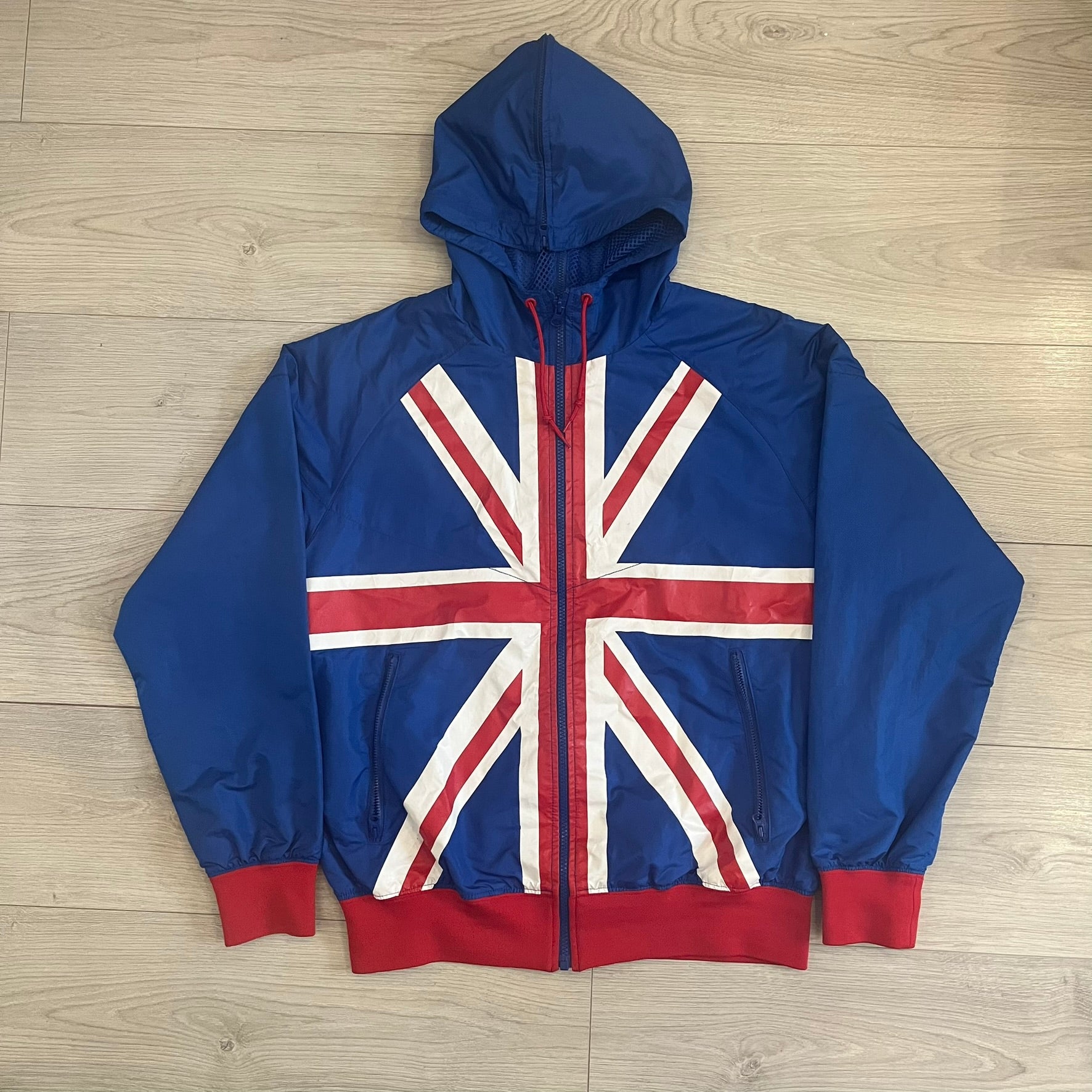 Nike UK Olympics 2008 Split Windbreaker designed by Jarrett Reynolds. Features a blue base with Union Jack-inspired red and white stripes on the front, representing Great Britain. Dual side zippers allow the jacket to be split in half and combined with other pieces from the collection, with a subtle United Kingdom flags inside the pocket.