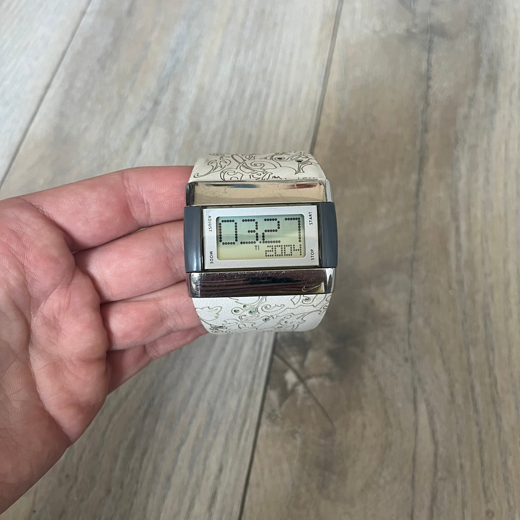 Nike WC0033 Merge Transit Digital Watch is a digital wristwatch featuring a silver-tone stainless steel case with a digital display and two gray side buttons on each side. The strap is white with engraved floral-style graphics on the outer side and a grey inner lining, made from a combination of leather, plastic, and rubber materials. The watch includes multiple modes and is water resistant up to 50 meters.
