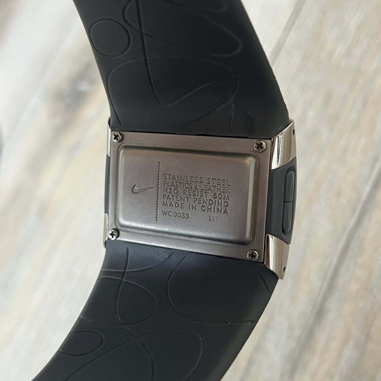 Nike WC0033 Merge Transit Digital Watch is a digital wristwatch featuring a silver-tone stainless steel case with a digital display and two gray side buttons on each side. The strap is white with engraved floral-style graphics on the outer side and a grey inner lining, made from a combination of leather, plastic, and rubber materials. The watch includes multiple modes and is water resistant up to 50 meters.
