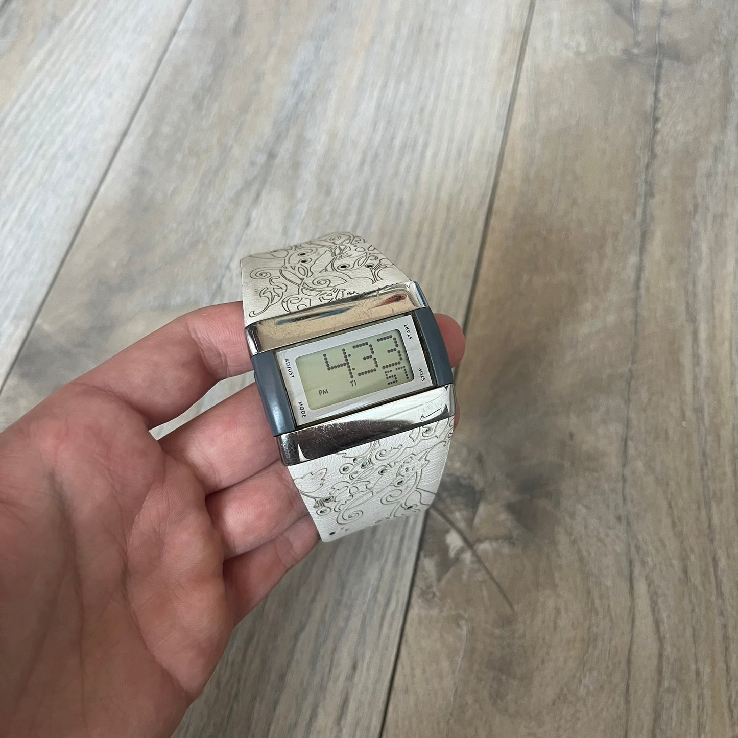 Nike WC0033 Merge Transit Digital Watch is a digital wristwatch featuring a silver-tone stainless steel case with a digital display and two gray side buttons on each side. The strap is white with engraved floral-style graphics on the outer side and a grey inner lining, made from a combination of leather, plastic, and rubber materials. The watch includes multiple modes and is water resistant up to 50 meters.

