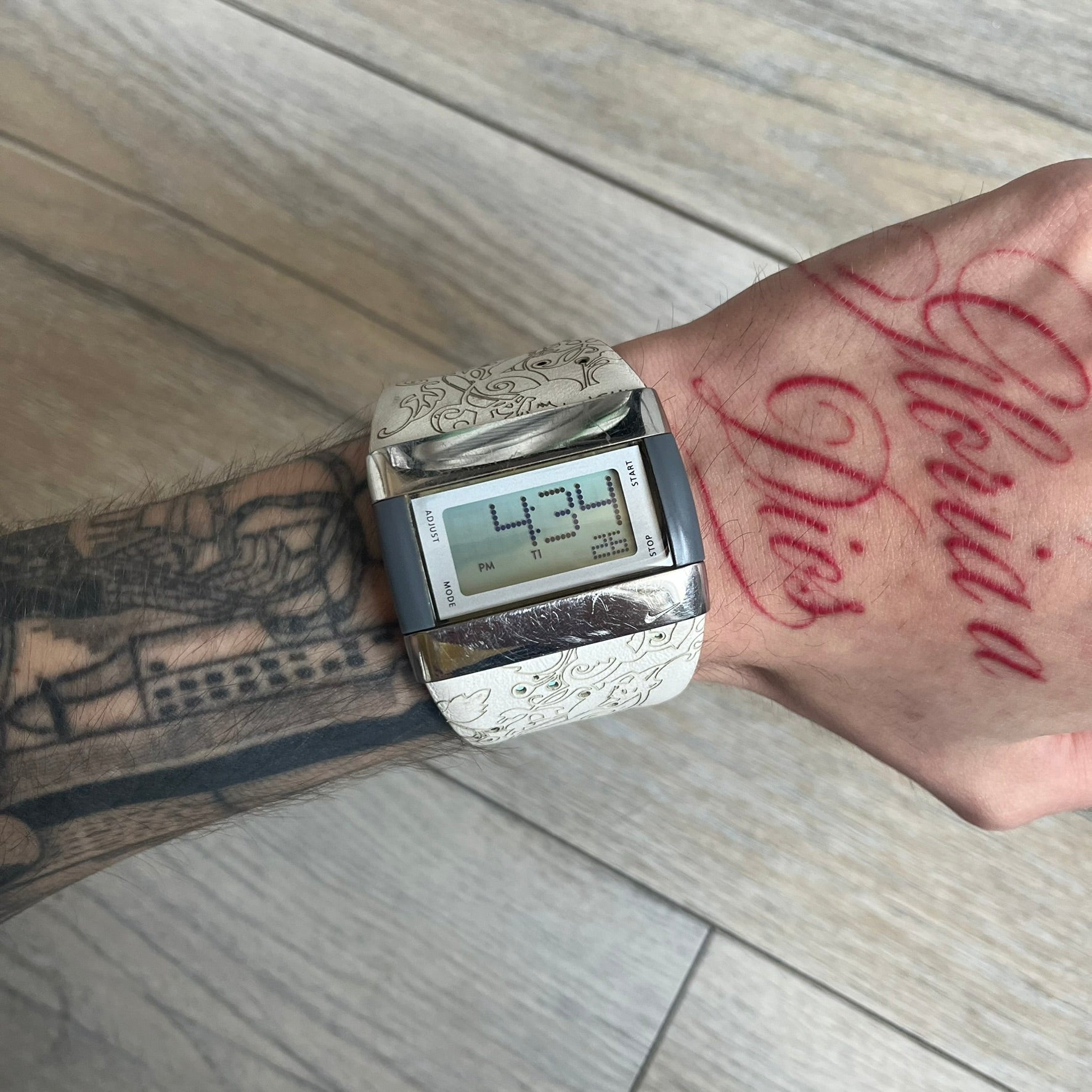 Nike WC0033 Merge Transit Digital Watch is a digital wristwatch featuring a silver-tone stainless steel case with a digital display and two gray side buttons on each side. The strap is white with engraved floral-style graphics on the outer side and a grey inner lining, made from a combination of leather, plastic, and rubber materials. The watch includes multiple modes and is water resistant up to 50 meters.
