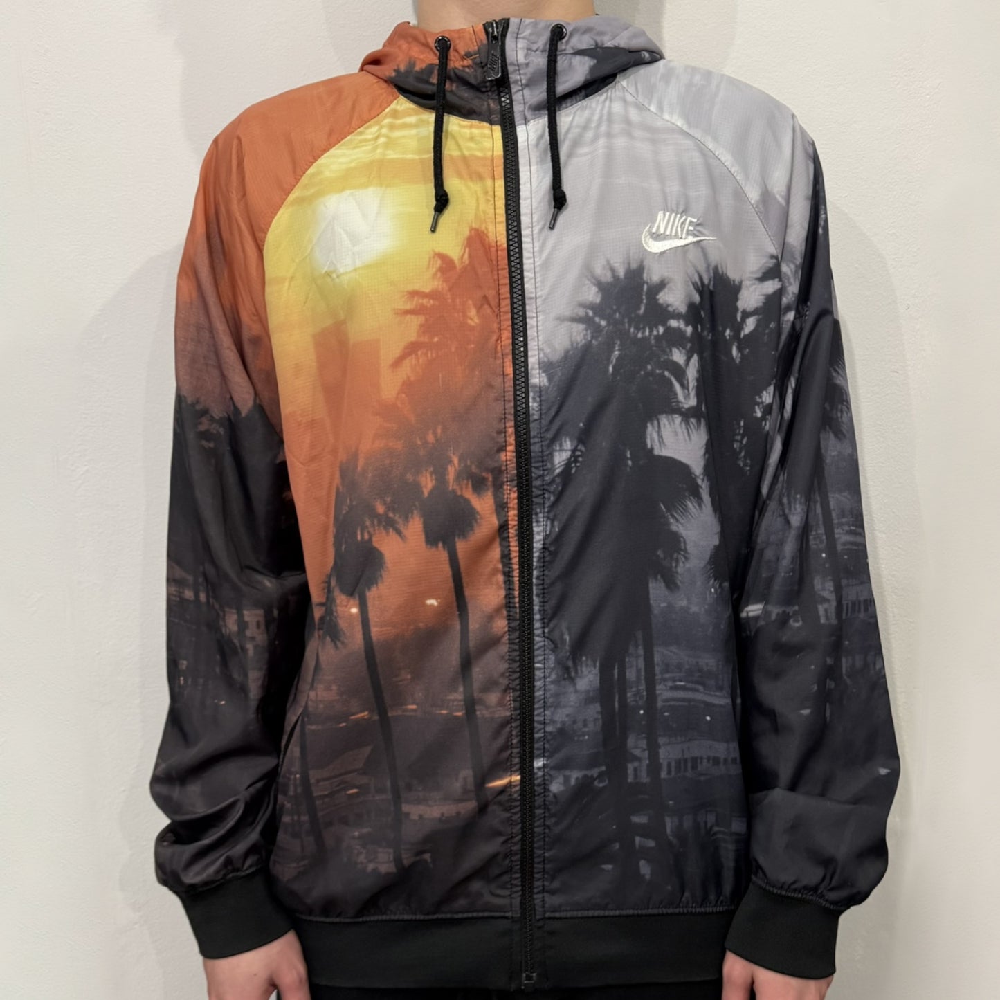 This Nike “Beach Sunset” Split Windbreaker merges two distinct colorways into one. Created from dual jackets that zip together through the front and back. The all-over print features palms, clouds, and a vibrant sunset, with one side in yellow–orange tones and the opposite in grey with black palm silhouettes. A minimal white Swoosh embroidery adds the final touch to this rare and highly collectible windbreaker.
