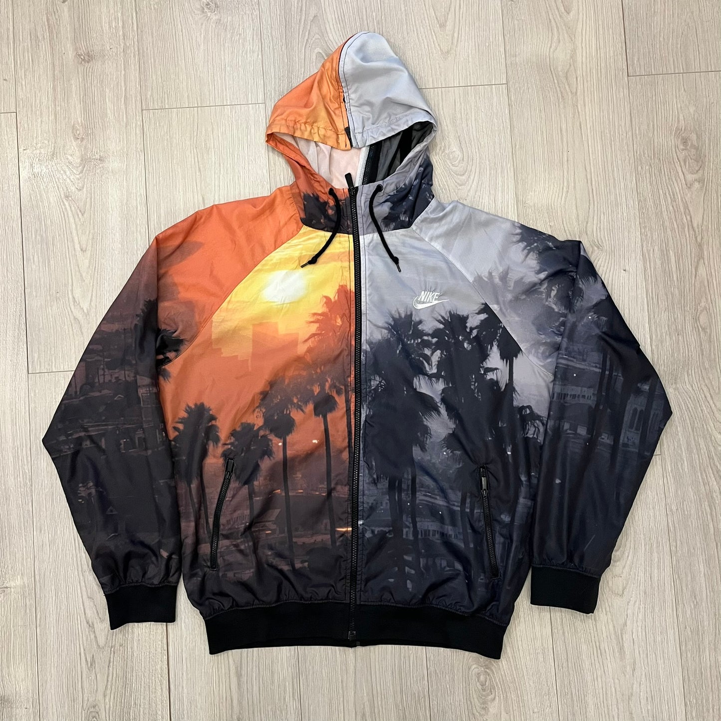 This Nike “Beach Sunset” Split Windbreaker merges two distinct colorways into one. Created from dual jackets that zip together through the front and back. The all-over print features palms, clouds, and a vibrant sunset, with one side in yellow–orange tones and the opposite in grey with black palm silhouettes. A minimal white Swoosh embroidery adds the final touch to this rare and highly collectible windbreaker.