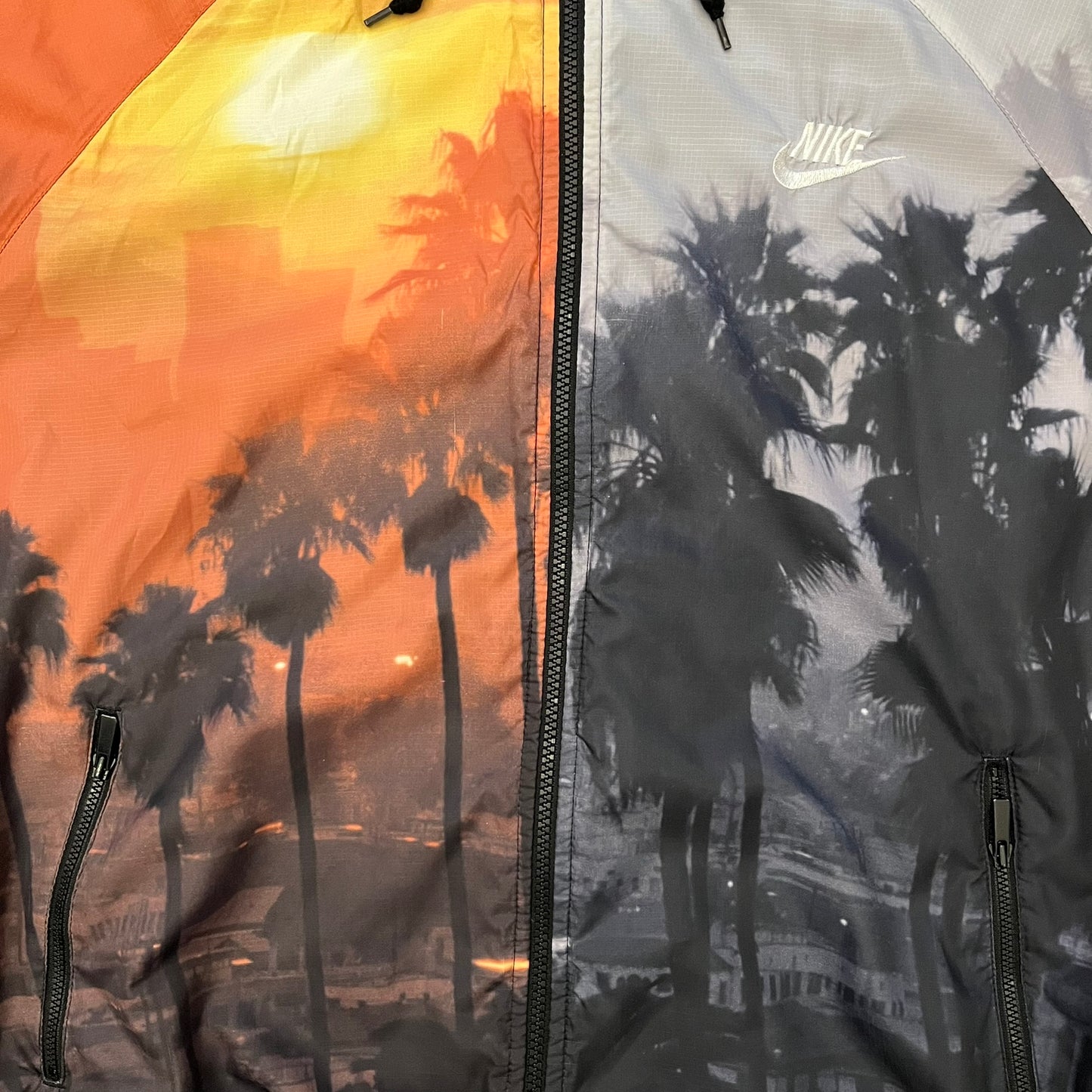 This Nike “Beach Sunset” Split Windbreaker merges two distinct colorways into one. Created from dual jackets that zip together through the front and back. The all-over print features palms, clouds, and a vibrant sunset, with one side in yellow–orange tones and the opposite in grey with black palm silhouettes. A minimal white Swoosh embroidery adds the final touch to this rare and highly collectible windbreaker.