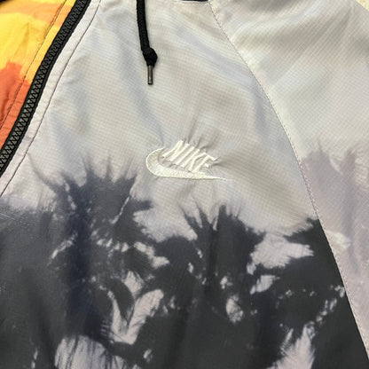 This Nike “Beach Sunset” Split Windbreaker merges two distinct colorways into one. Created from dual jackets that zip together through the front and back. The all-over print features palms, clouds, and a vibrant sunset, with one side in yellow–orange tones and the opposite in grey with black palm silhouettes. A minimal white Swoosh embroidery adds the final touch to this rare and highly collectible windbreaker.
