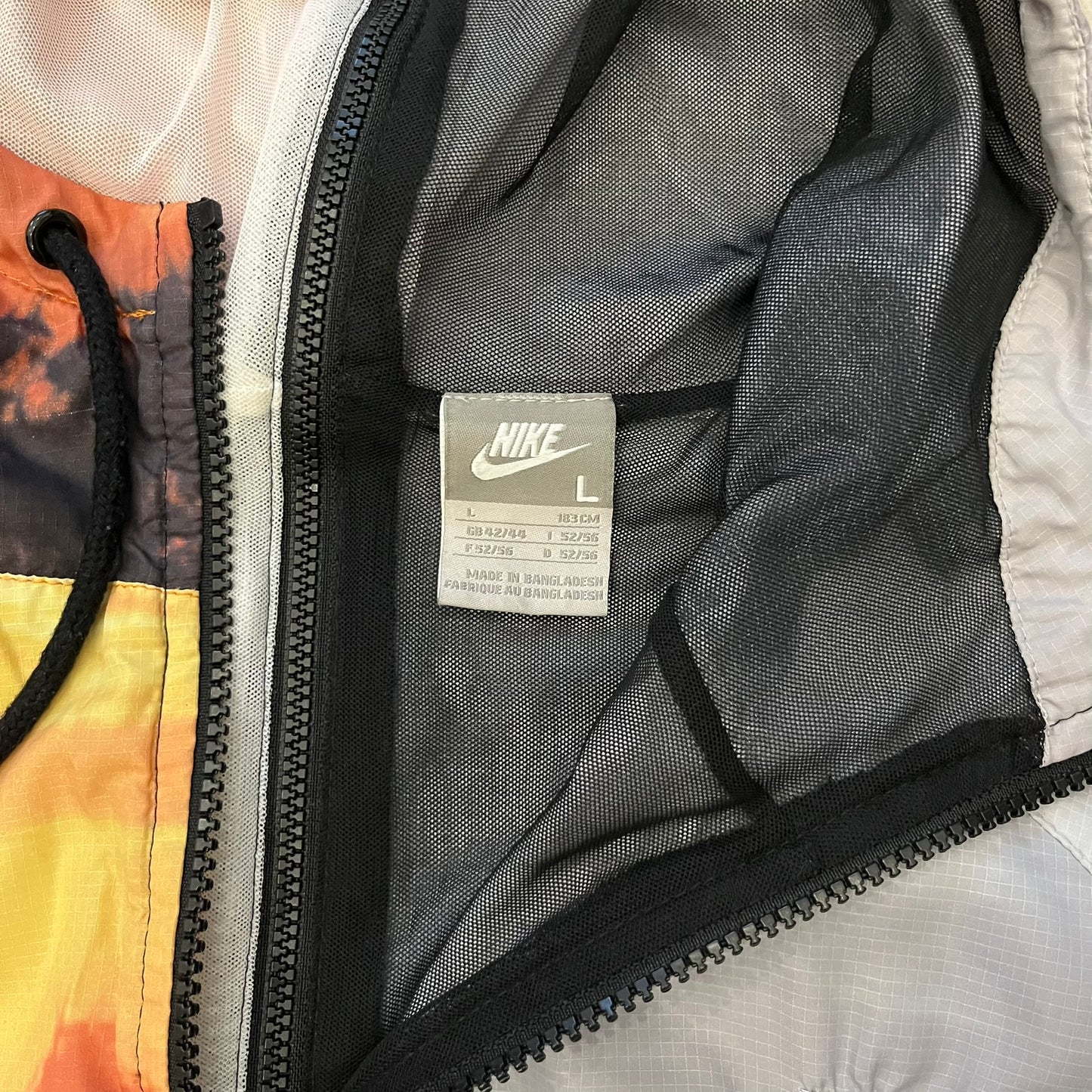 This Nike “Beach Sunset” Split Windbreaker merges two distinct colorways into one. Created from dual jackets that zip together through the front and back. The all-over print features palms, clouds, and a vibrant sunset, with one side in yellow–orange tones and the opposite in grey with black palm silhouettes. A minimal white Swoosh embroidery adds the final touch to this rare and highly collectible windbreaker.