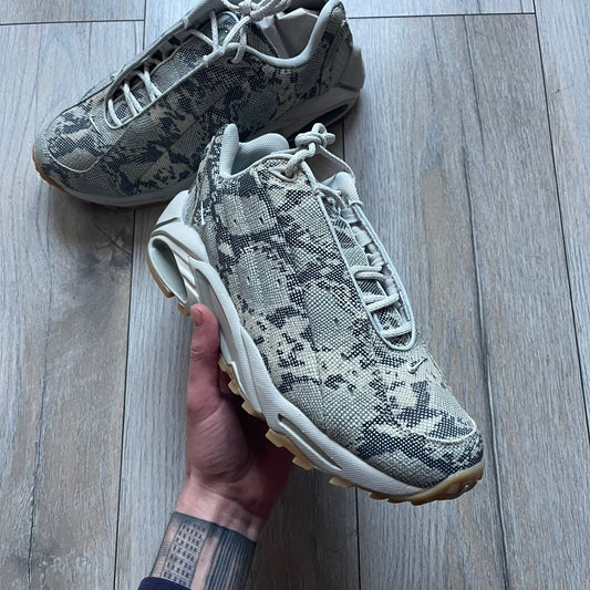 The Nike x NOCTA Hot Step Air Terra “Snakeskin” Light Bone Sneakers feature a low-top silhouette with a textured faux snakeskin upper in Light Bone tones mixed with cobblestone and gum light brown accents. The shoe includes small Swoosh logos on the sides, NOCTA three-star branding on the tongue, perforated quarter panels, and a metallic heel tag with Gore-Tex embossing. Finished with a white midsole housing a visible Air unit at the heel and a gum rubber outsole, style code DR0508-001, colorway Light Bone/