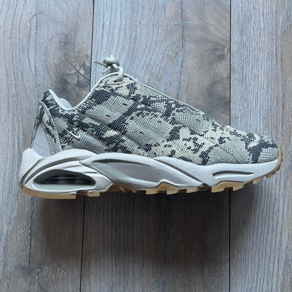 The Nike x NOCTA Hot Step Air Terra “Snakeskin” Light Bone Sneakers feature a low-top silhouette with a textured faux snakeskin upper in Light Bone tones mixed with cobblestone and gum light brown accents. The shoe includes small Swoosh logos on the sides, NOCTA three-star branding on the tongue, perforated quarter panels, and a metallic heel tag with Gore-Tex embossing. Finished with a white midsole housing a visible Air unit at the heel and a gum rubber outsole, style code DR0508-001, colorway Light Bone/