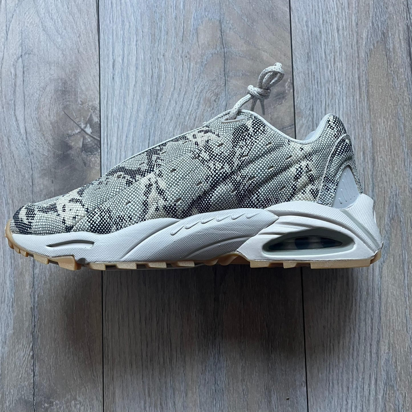 The Nike x NOCTA Hot Step Air Terra “Snakeskin” Light Bone Sneakers feature a low-top silhouette with a textured faux snakeskin upper in Light Bone tones mixed with cobblestone and gum light brown accents. The shoe includes small Swoosh logos on the sides, NOCTA three-star branding on the tongue, perforated quarter panels, and a metallic heel tag with Gore-Tex embossing. Finished with a white midsole housing a visible Air unit at the heel and a gum rubber outsole, style code DR0508-001, colorway Light Bone/