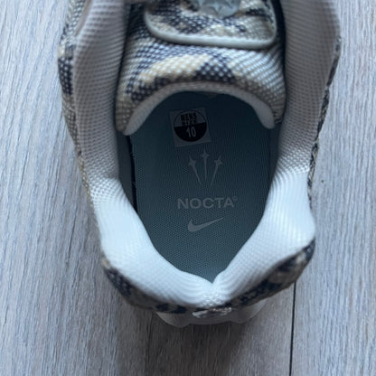 The Nike x NOCTA Hot Step Air Terra “Snakeskin” Light Bone Sneakers feature a low-top silhouette with a textured faux snakeskin upper in Light Bone tones mixed with cobblestone and gum light brown accents. The shoe includes small Swoosh logos on the sides, NOCTA three-star branding on the tongue, perforated quarter panels, and a metallic heel tag with Gore-Tex embossing. Finished with a white midsole housing a visible Air unit at the heel and a gum rubber outsole, style code DR0508-001, colorway Light Bone/