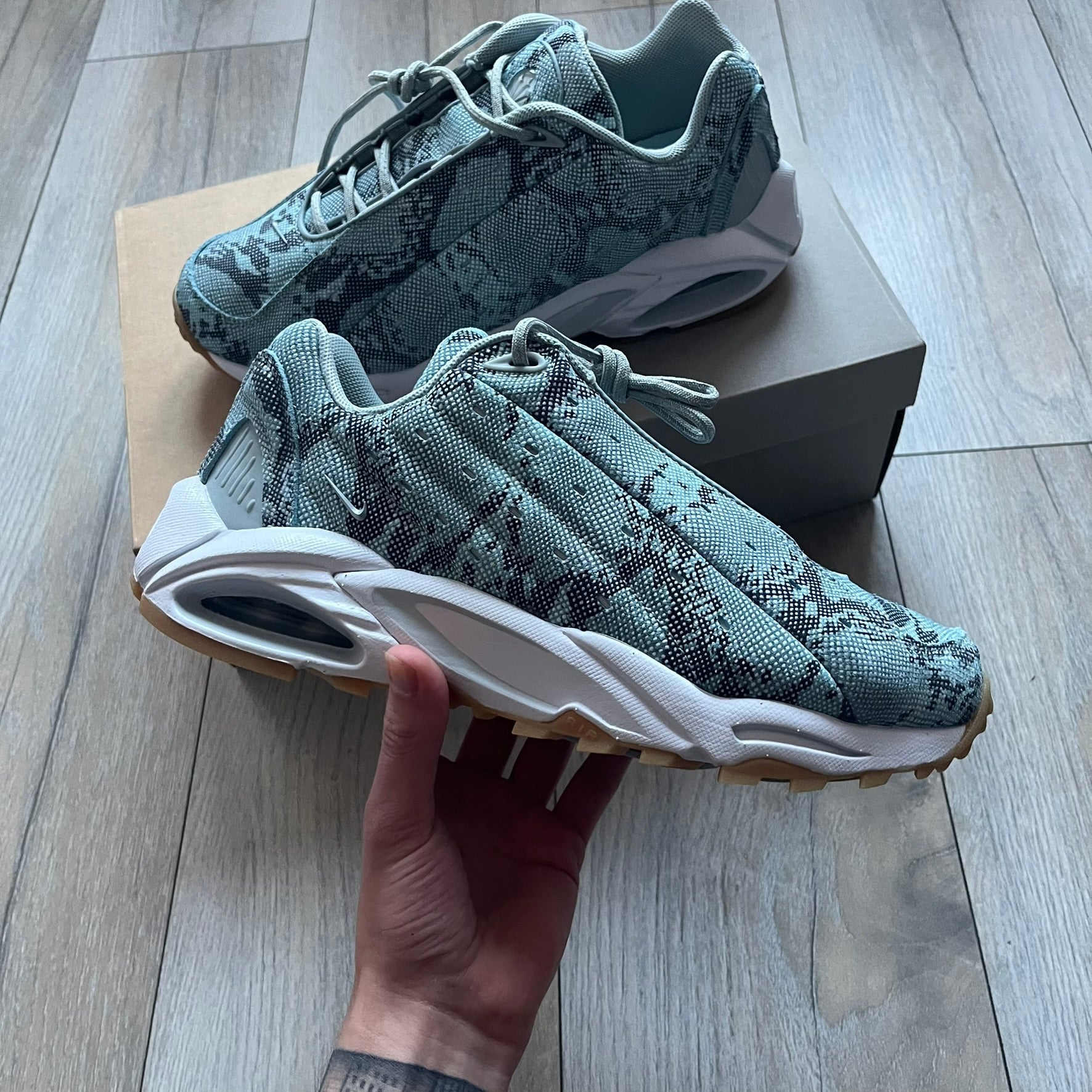 The Nike x NOCTA Hot Step Air Terra “Snakeskin” Light Pumice Sneakers feature a low-top build with a faux snakeskin-textured upper in light pumice blue and grey tones. Details include small 3D mini Swooshes on the sides, reflective piping around the eyestay, oval perforations at the midfoot with a reflective underlay, and NOCTA branding on the tongue and insole. Finished with a white foam midsole inspired by the Air Terra Humara with Zoom Air in the forefoot, a visible Max Air unit in the heel, and a gum ru