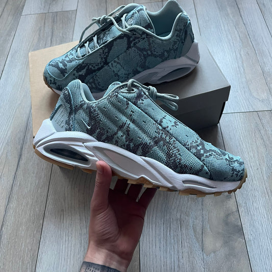 The Nike x NOCTA Hot Step Air Terra “Snakeskin” Light Pumice Sneakers feature a low-top build with a faux snakeskin-textured upper in light pumice blue and grey tones. Details include small 3D mini Swooshes on the sides, reflective piping around the eyestay, oval perforations at the midfoot with a reflective underlay, and NOCTA branding on the tongue and insole. Finished with a white foam midsole inspired by the Air Terra Humara with Zoom Air in the forefoot, a visible Max Air unit in the heel, and a gum ru