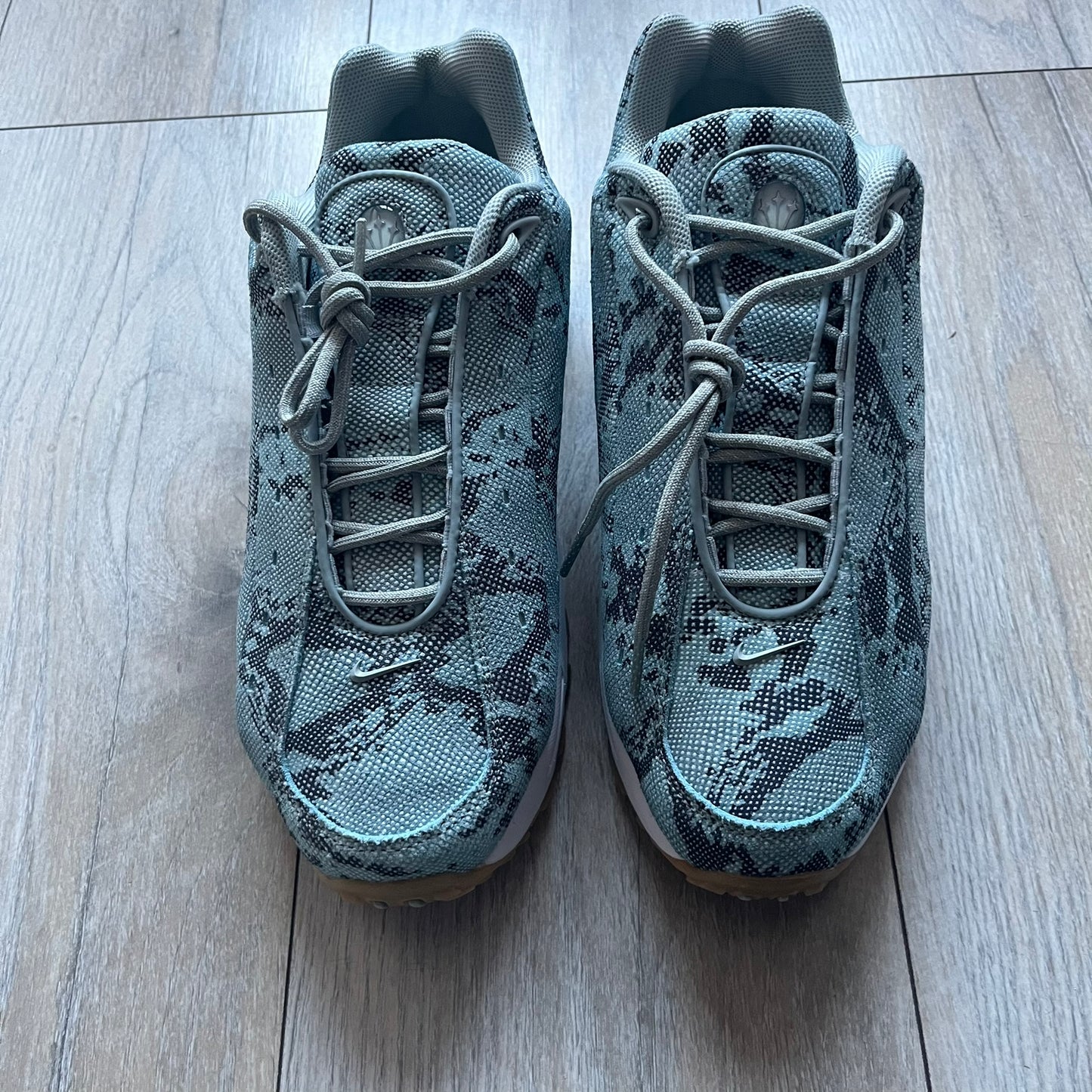 The Nike x NOCTA Hot Step Air Terra “Snakeskin” Light Pumice Sneakers feature a low-top build with a faux snakeskin-textured upper in light pumice blue and grey tones. Details include small 3D mini Swooshes on the sides, reflective piping around the eyestay, oval perforations at the midfoot with a reflective underlay, and NOCTA branding on the tongue and insole. Finished with a white foam midsole inspired by the Air Terra Humara with Zoom Air in the forefoot, a visible Max Air unit in the heel, and a gum ru