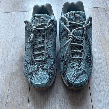 The Nike x NOCTA Hot Step Air Terra “Snakeskin” Light Pumice Sneakers feature a low-top build with a faux snakeskin-textured upper in light pumice blue and grey tones. Details include small 3D mini Swooshes on the sides, reflective piping around the eyestay, oval perforations at the midfoot with a reflective underlay, and NOCTA branding on the tongue and insole. Finished with a white foam midsole inspired by the Air Terra Humara with Zoom Air in the forefoot, a visible Max Air unit in the heel, and a gum ru