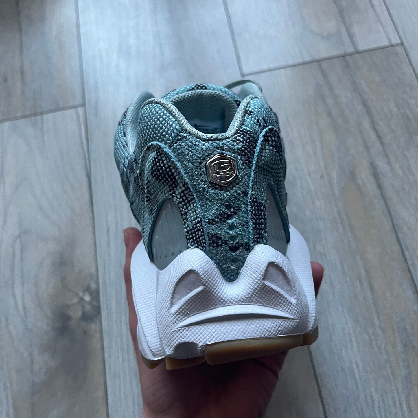 The Nike x NOCTA Hot Step Air Terra “Snakeskin” Light Pumice Sneakers feature a low-top build with a faux snakeskin-textured upper in light pumice blue and grey tones. Details include small 3D mini Swooshes on the sides, reflective piping around the eyestay, oval perforations at the midfoot with a reflective underlay, and NOCTA branding on the tongue and insole. Finished with a white foam midsole inspired by the Air Terra Humara with Zoom Air in the forefoot, a visible Max Air unit in the heel, and a gum ru