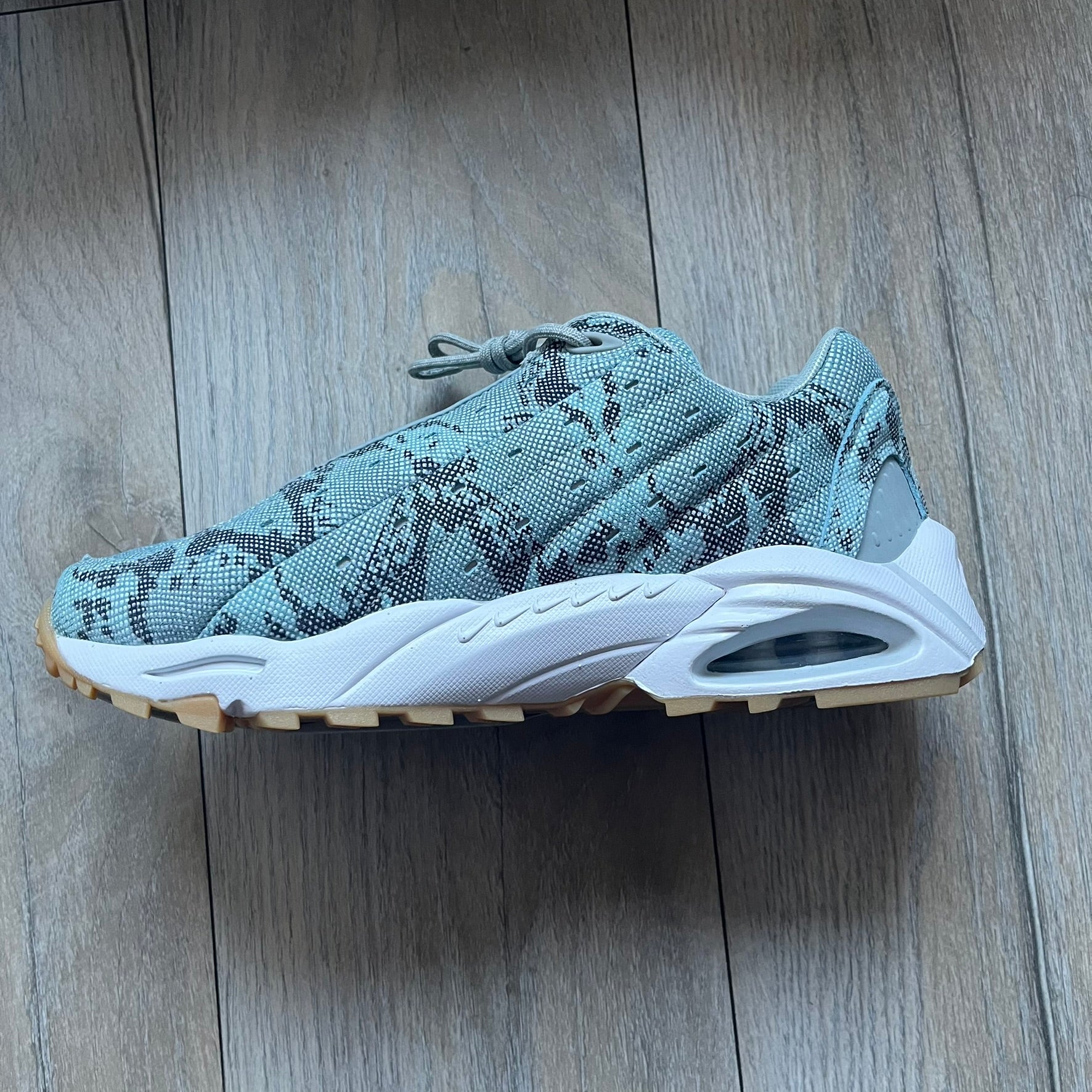 The Nike x NOCTA Hot Step Air Terra “Snakeskin” Light Pumice Sneakers feature a low-top build with a faux snakeskin-textured upper in light pumice blue and grey tones. Details include small 3D mini Swooshes on the sides, reflective piping around the eyestay, oval perforations at the midfoot with a reflective underlay, and NOCTA branding on the tongue and insole. Finished with a white foam midsole inspired by the Air Terra Humara with Zoom Air in the forefoot, a visible Max Air unit in the heel, and a gum ru