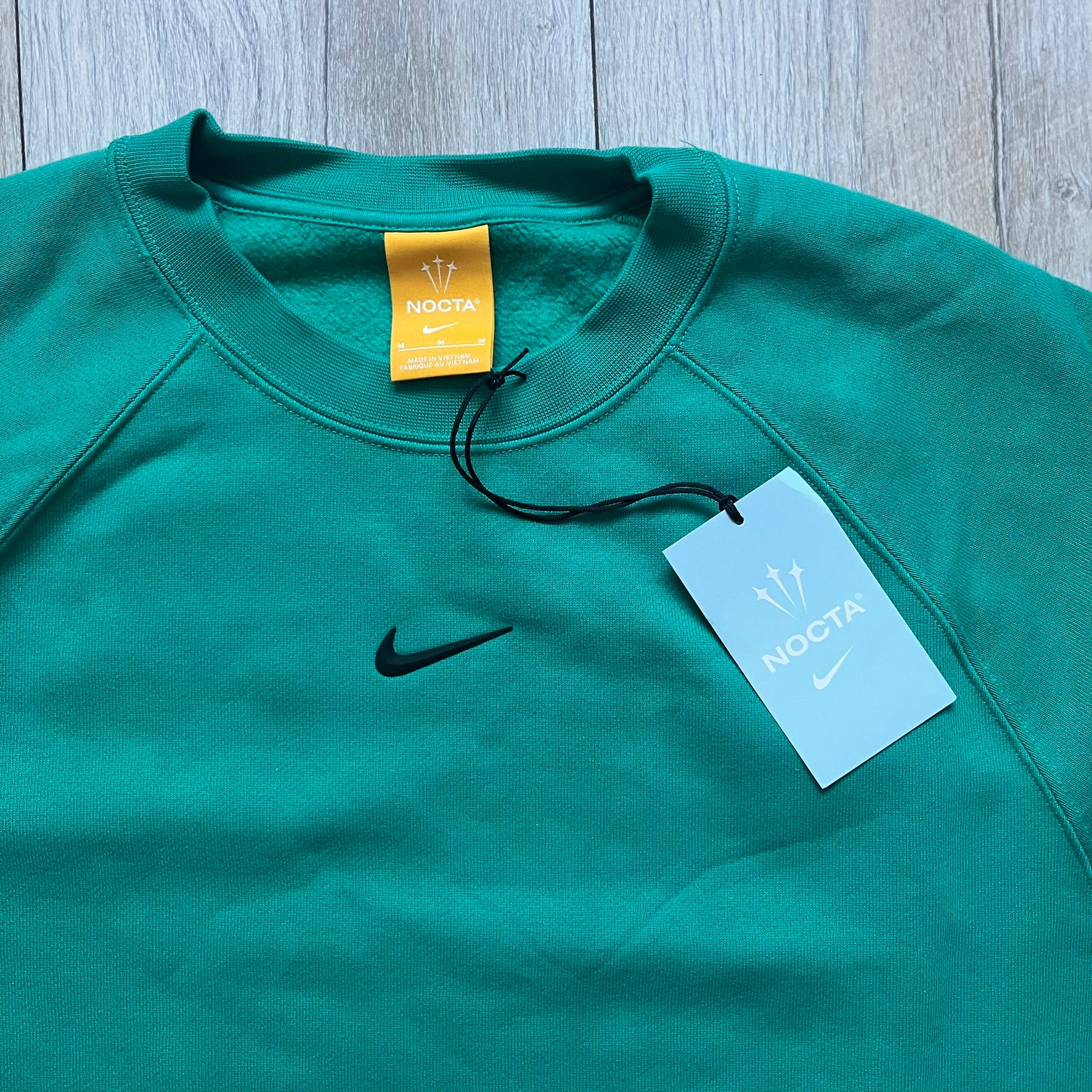 Nike x NOCTA Fleece “Neptune Green” Crewneck Sweatshirt FN8160-370 is a men’s pullover from the Cardinal Stock collection, finished in a Neptune Green/Black colorway. It features a regular fit with a round neckline, long sleeves, elastic cuffs, and an elasticated lower hem.&nbsp;Made from 82% cotton and 18% polyester fleece, the sweatshirt includes a silicone Swoosh logo and signature NOCTA branding, along with a back print, sleeve print, and 3D logo detailing. Made in Vietnam.