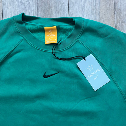 Nike x NOCTA Fleece “Neptune Green” Crewneck Sweatshirt FN8160-370 is a men’s pullover from the Cardinal Stock collection, finished in a Neptune Green/Black colorway. It features a regular fit with a round neckline, long sleeves, elastic cuffs, and an elasticated lower hem.&nbsp;Made from 82% cotton and 18% polyester fleece, the sweatshirt includes a silicone Swoosh logo and signature NOCTA branding, along with a back print, sleeve print, and 3D logo detailing. Made in Vietnam.