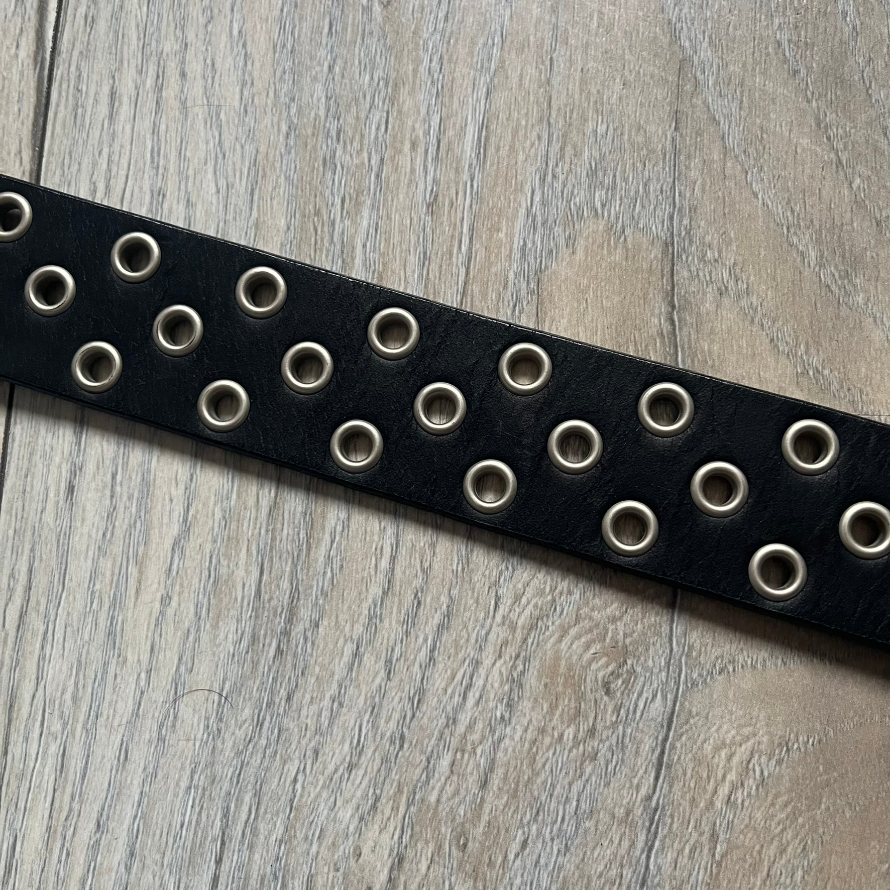 Oakley 2004 Black Leather Skull Belt is a genuine black leather belt featuring full-length holes for adjustable sizing. The belt end is embossed with a carved Oakley logo, and it includes a metal skull buckle with Oakley branding. Made in 2004, it combines durable leather construction with signature Oakley design details.