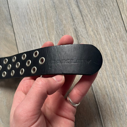 Oakley 2004 Black Leather Skull Belt is a genuine black leather belt featuring full-length holes for adjustable sizing. The belt end is embossed with a carved Oakley logo, and it includes a metal skull buckle with Oakley branding. Made in 2004, it combines durable leather construction with signature Oakley design details.