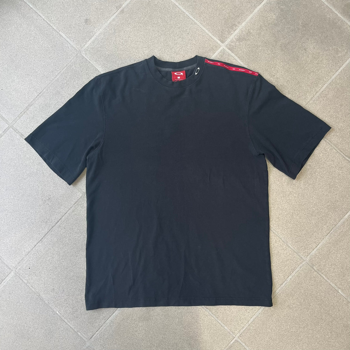 Oakley 2000's Metal Ellipse Black Tee is a black T-shirt made from 95% cotton and 5% spandex, featuring a raised collar with a metal Oakley Ellipse logo and a red stripe printed with Oakley logos on one shoulder.