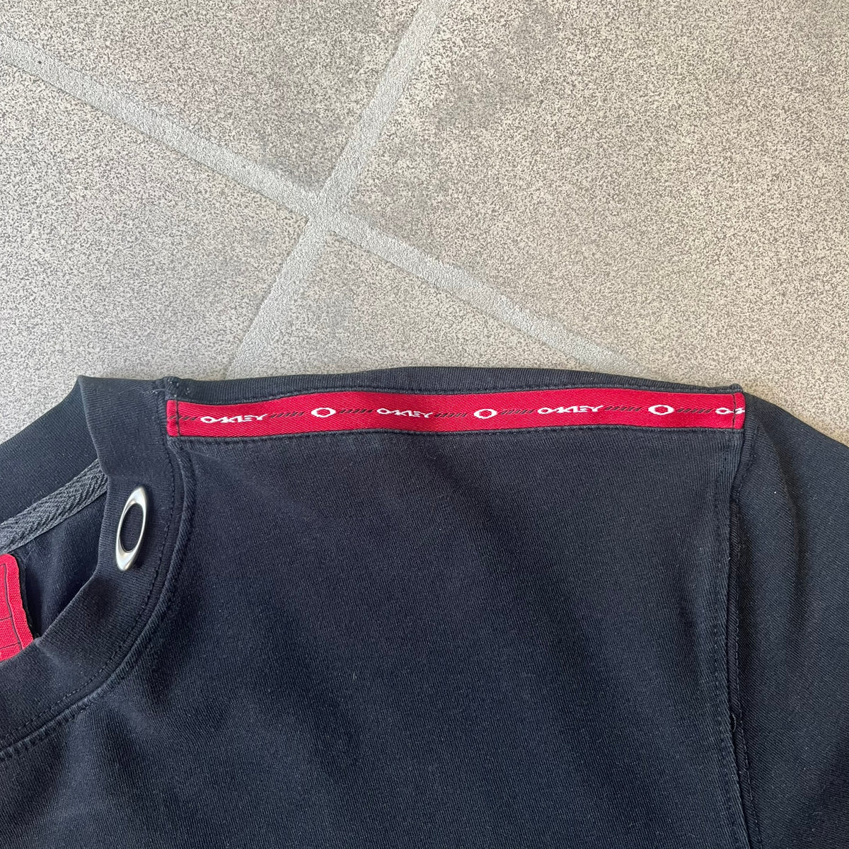 Oakley 2000's Metal Ellipse Black Tee is a black T-shirt made from 95% cotton and 5% spandex, featuring a raised collar with a metal Oakley Ellipse logo and a red stripe printed with Oakley logos on one shoulder.