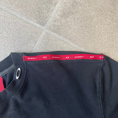 Oakley 2000's Metal Ellipse Black Tee is a black T-shirt made from 95% cotton and 5% spandex, featuring a raised collar with a metal Oakley Ellipse logo and a red stripe printed with Oakley logos on one shoulder.