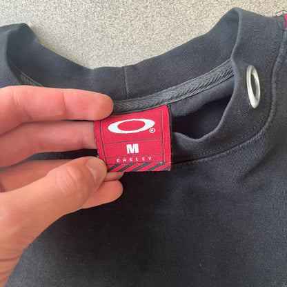 Oakley 2000's Metal Ellipse Black Tee is a black T-shirt made from 95% cotton and 5% spandex, featuring a raised collar with a metal Oakley Ellipse logo and a red stripe printed with Oakley logos on one shoulder.