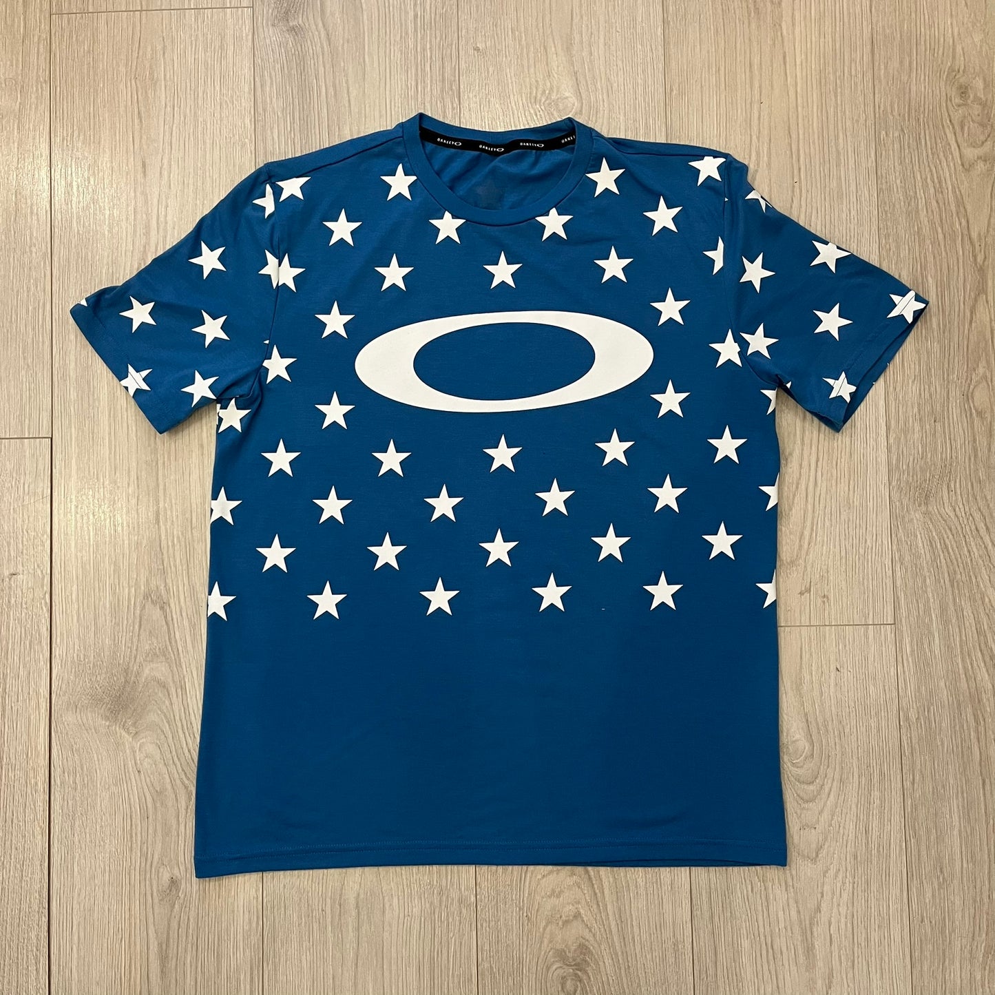 Oakley Crazy Stars Tee in blue with an all-over white star print covering the top two-thirds of the front, back, and sleeves. Features a large Oakley “O” logo on the front. Made from a cotton/polyester blend in El Salvador.