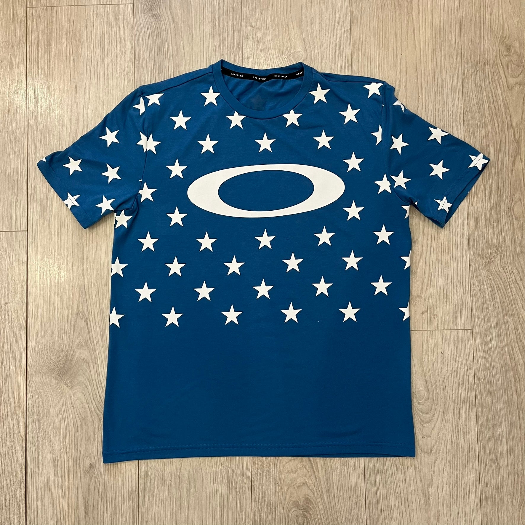 Oakley Crazy Stars Tee in blue with an all-over white star print covering the top two-thirds of the front, back, and sleeves. Features a large Oakley “O” logo on the front. Made from a cotton/polyester blend in El Salvador.