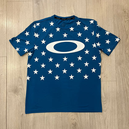 Oakley Crazy Stars Tee in blue with an all-over white star print covering the top two-thirds of the front, back, and sleeves. Features a large Oakley “O” logo on the front. Made from a cotton/polyester blend in El Salvador.