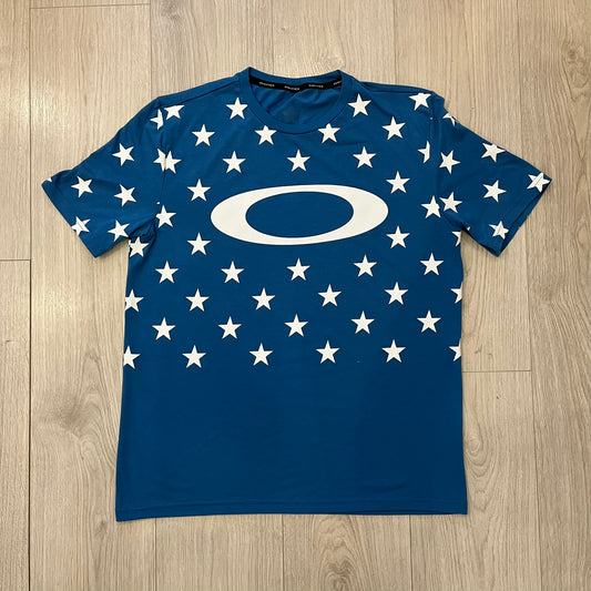 Oakley Crazy Stars Tee in blue with an all-over white star print covering the top two-thirds of the front, back, and sleeves. Features a large Oakley “O” logo on the front. Made from a cotton/polyester blend in El Salvador.