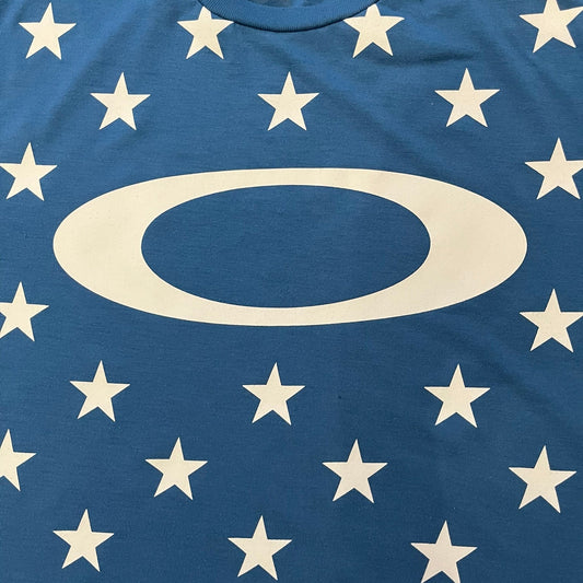 Oakley Crazy Stars Tee in blue with an all-over white star print covering the top two-thirds of the front, back, and sleeves. Features a large Oakley “O” logo on the front. Made from a cotton/polyester blend in El Salvador.