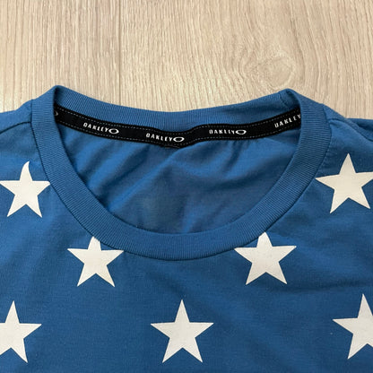 Oakley Crazy Stars Tee in blue with an all-over white star print covering the top two-thirds of the front, back, and sleeves. Features a large Oakley “O” logo on the front. Made from a cotton/polyester blend in El Salvador.