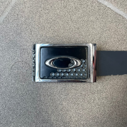 Oakley Ellipse 2010's Gray Leather Belt features a gray leather strap with a cut-to-size construction for adjustable length. The metal rectangular buckle has a silver outer frame, black center insert, and a silver Oakley Ellipse logo in the middle, with an engraved Oakley logo on the side.