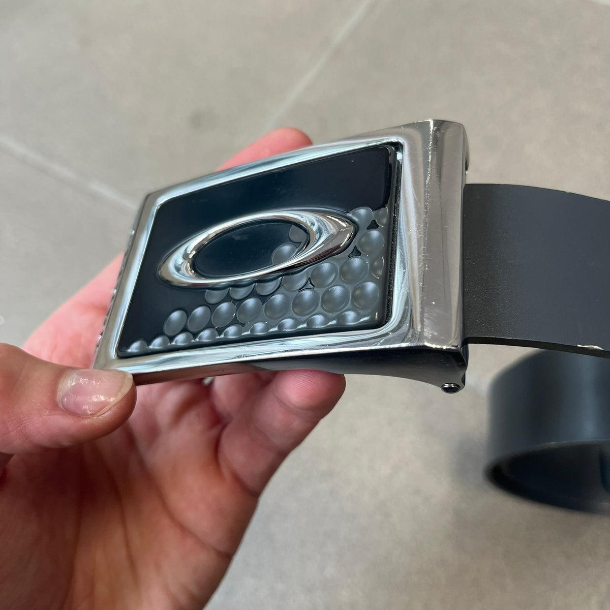 Oakley Ellipse 2010's Gray Leather Belt features a gray leather strap with a cut-to-size construction for adjustable length. The metal rectangular buckle has a silver outer frame, black center insert, and a silver Oakley Ellipse logo in the middle, with an engraved Oakley logo on the side.