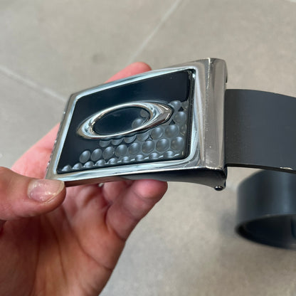 Oakley Ellipse 2010's Gray Leather Belt features a gray leather strap with a cut-to-size construction for adjustable length. The metal rectangular buckle has a silver outer frame, black center insert, and a silver Oakley Ellipse logo in the middle, with an engraved Oakley logo on the side.