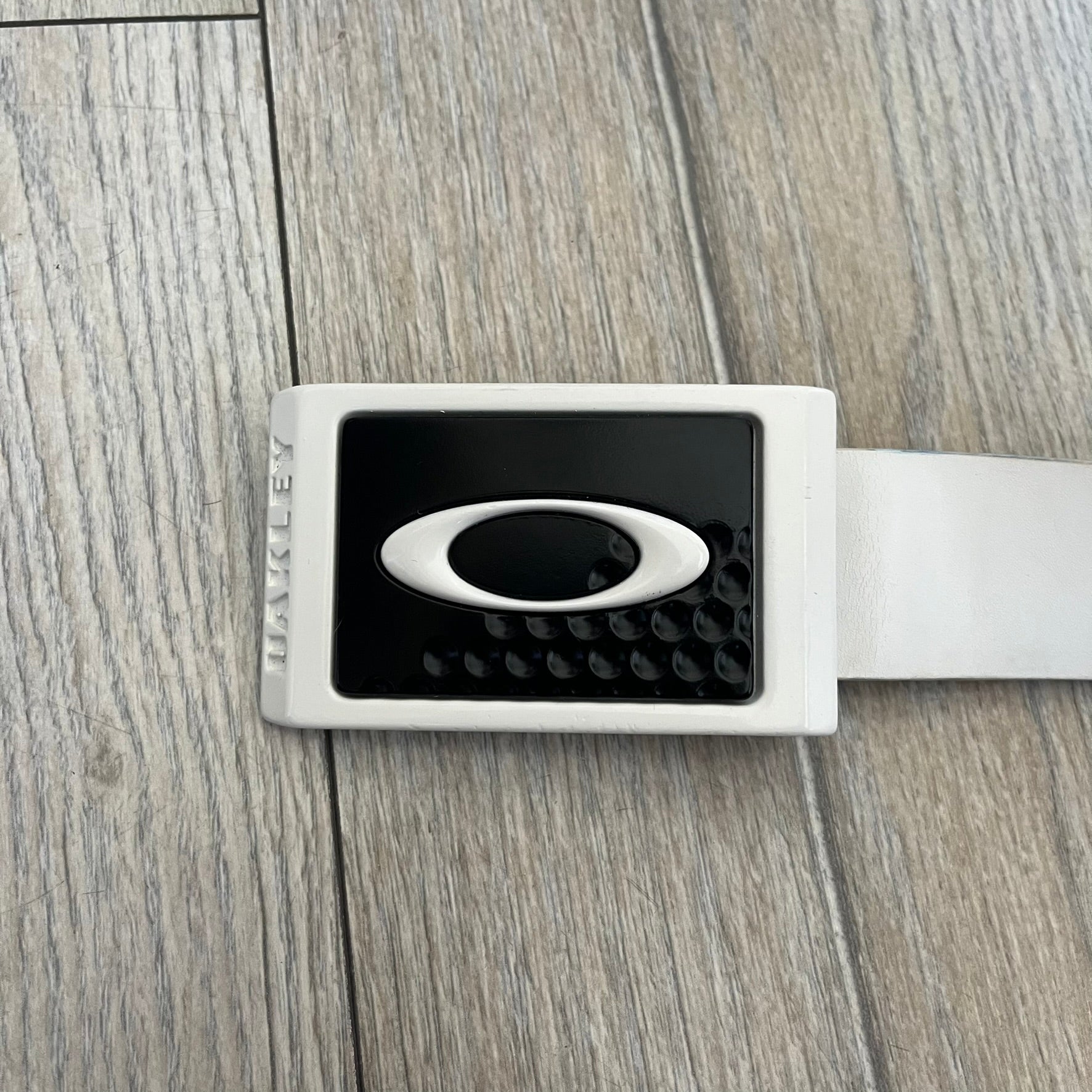 Oakley Ellipse 2010s White Leather Belt features a white leather strap with a cut-to-size construction for adjustable length. The metal rectangular buckle has a white outer frame, black center insert, and an Oakley Ellipse logo in the middle, with an engraved Oakley logo on the side. The set includes an additional replacement belt strap.