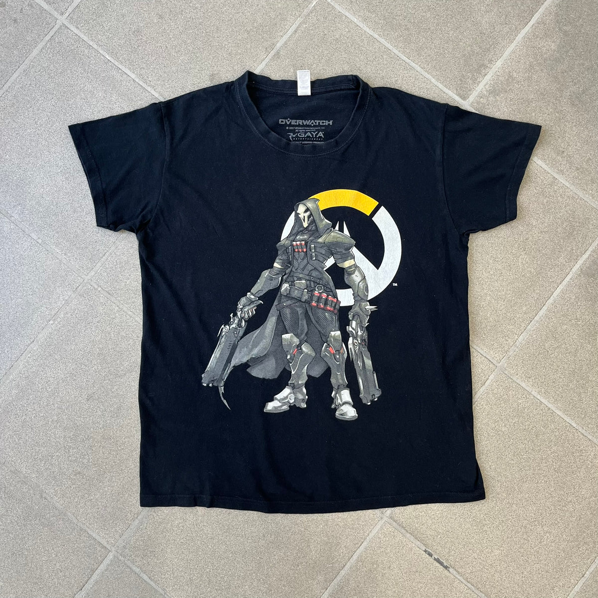 Overwatch 2017 Reaper Black Tee features a black construction with a full-color graphic of Reaper standing with his guns in front of the Overwatch logo, with a small “TM” mark beside it. Finished with a licensed 2017 Overwatch Gaya neck tag.
