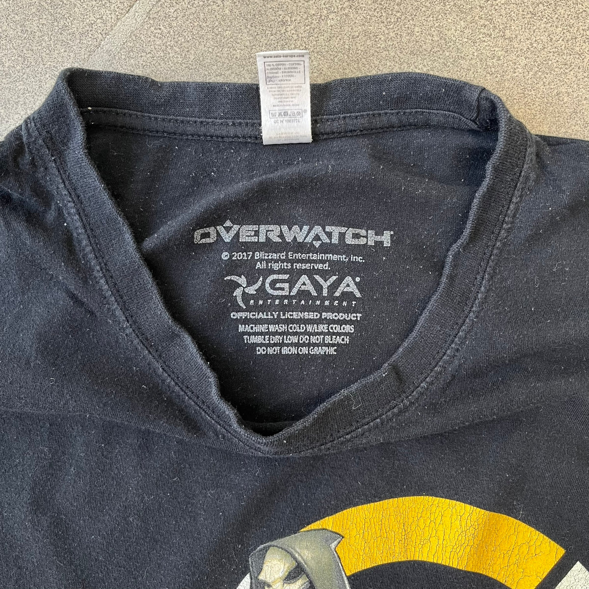 Overwatch 2017 Reaper Black Tee features a black construction with a full-color graphic of Reaper standing with his guns in front of the Overwatch logo, with a small “TM” mark beside it. Finished with a licensed 2017 Overwatch Gaya neck tag.
