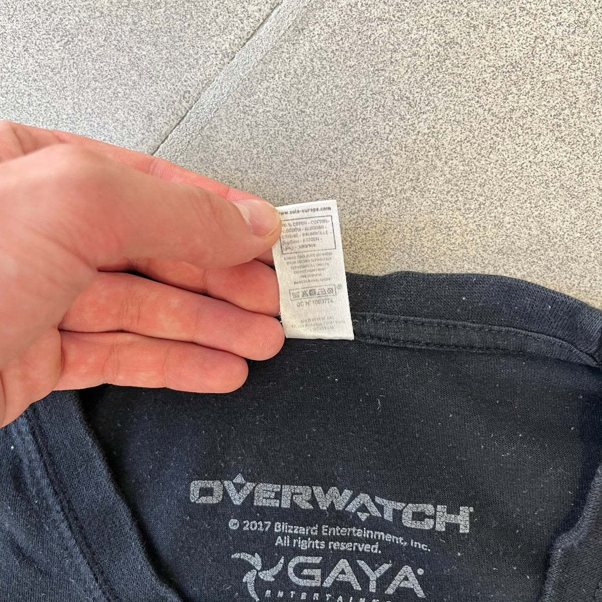 Overwatch 2017 Reaper Black Tee features a black construction with a full-color graphic of Reaper standing with his guns in front of the Overwatch logo, with a small “TM” mark beside it. Finished with a licensed 2017 Overwatch Gaya neck tag.
