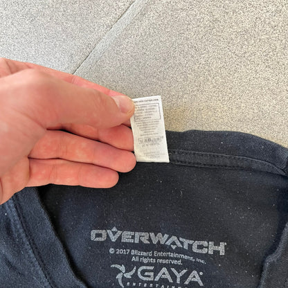 Overwatch 2017 Reaper Black Tee features a black construction with a full-color graphic of Reaper standing with his guns in front of the Overwatch logo, with a small “TM” mark beside it. Finished with a licensed 2017 Overwatch Gaya neck tag.
