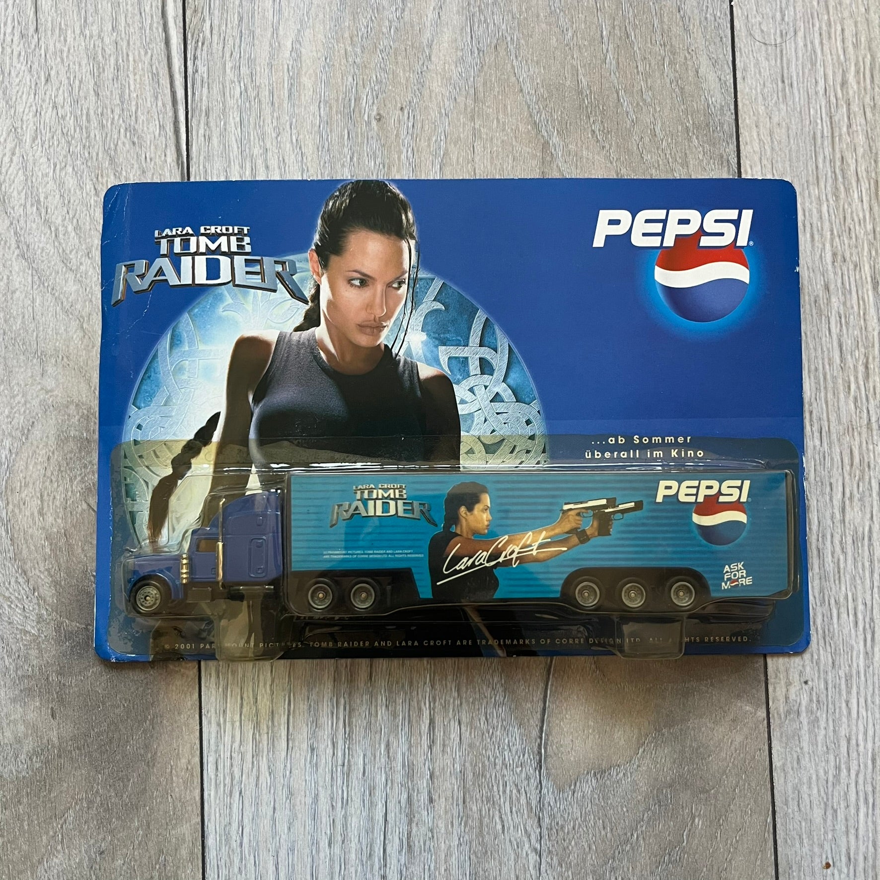 Pepsi 2001 Lara Croft Tomb Raider 1:87 Delivery Truck Model is a promotional die-cast style truck released alongside the film Lara Croft: Tomb Raider. The model features a blue Pepsi delivery truck with a large graphic of Lara Croft and Pepsi branding on the sides. It comes displayed in a clear plastic case attached to a printed cardboard backing with Tomb Raider and Pepsi graphics. The bottom of the case includes 2001 Paramount Pictures licensing.