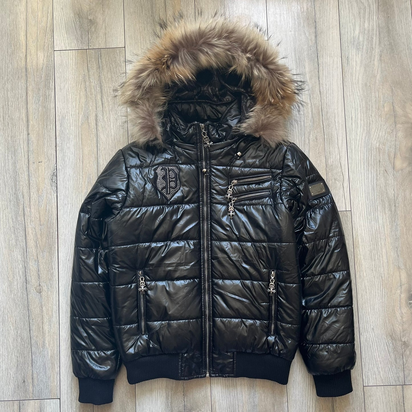 Philipp Plein 2010's Black Waxed Puffer Jacket is a black puffer jacket with a removable fur-trimmed hood attached by zipper. The jacket features heavy silver-tone hardware throughout, including large zipper pulls shaped as crosses with a skull in the center. The right chest has a crystal “P” patch set, while the left chest features two zip pockets. The hood drawstrings are finished with silver skull tips marked with a “P.”

One shoulder is finished with metal Philipp Plein logo plaque. The interior lining 